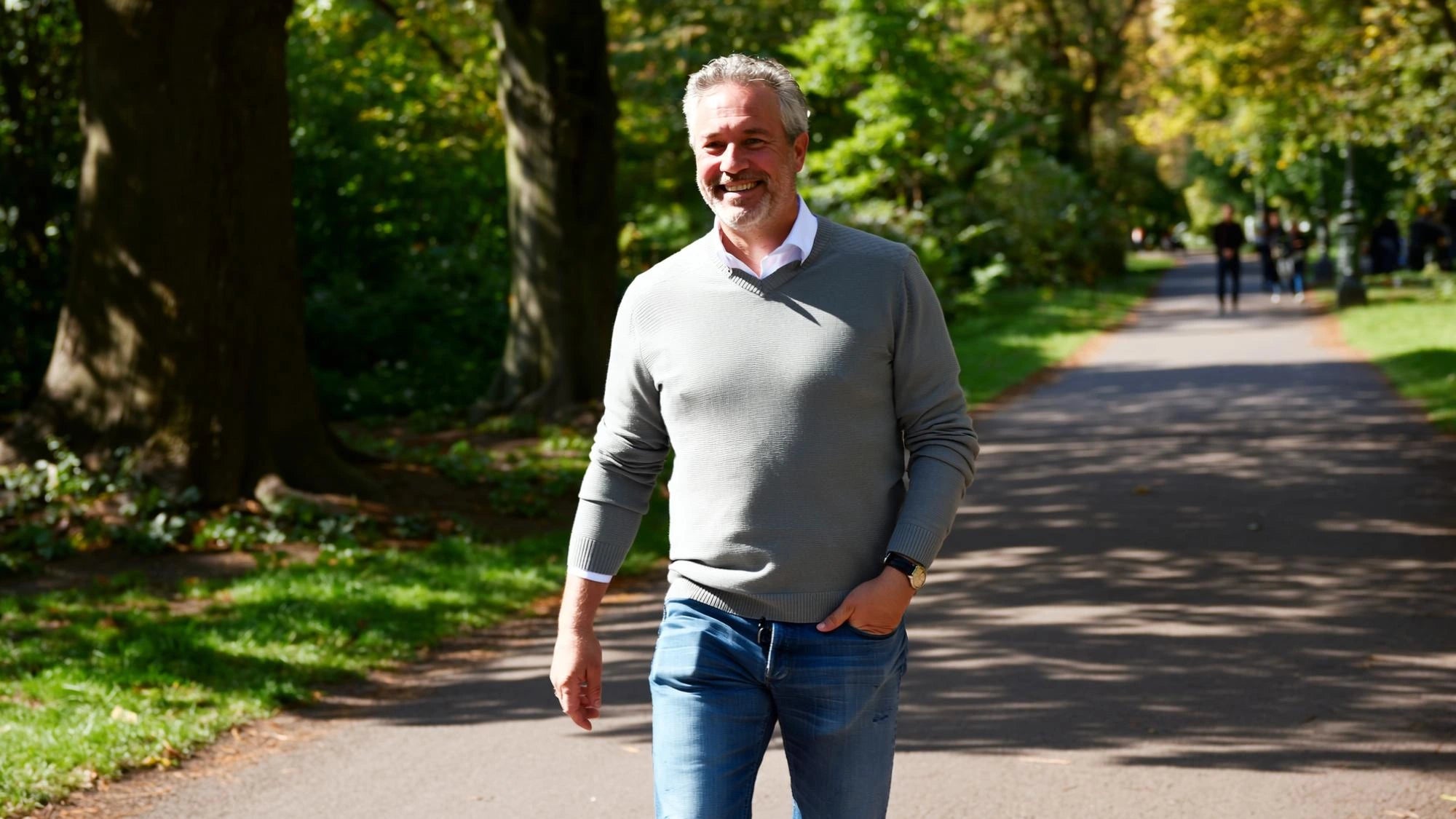 Man in his sixties walking at an easy pace along a tree-lined park path on a sunny morning